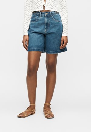 Kvinde iført mid-lange blå denimshorts, hvid cropped langærmet top og beige flade sandaler med stropper, stående mod en ensfarvet baggrund.