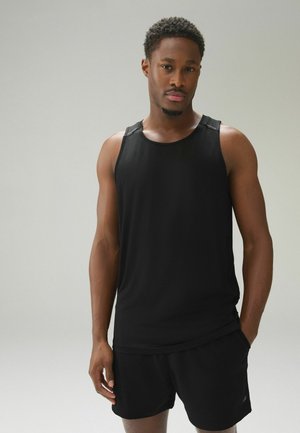 Mann mit kurzem lockigem Haar, der ein schwarzes ärmelloses Sportoberteil und schwarze Shorts trägt, vor einem schlichten hellen Hintergrund stehend.