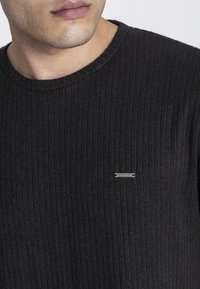 Čierny rebrovaný pletený sveter s okrúhlym výstrihom a striebornou štítkom na hrudi, zdôrazňujúci štruktúrovanú látku s vertikálnymi pruhmi.