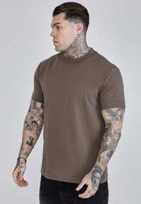 Ruda medvilnės marškinėliai su apvaliu kaklu ir trumpomis rankovėmis, dėvimi modelio, kurio rankos padengtos tatuiruotėmis, parodyti šoniniu profiliu lengvo fono atžvilgiu.