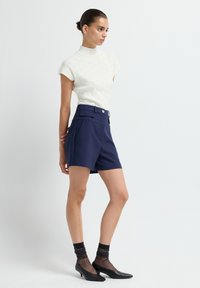 Vrouw staat in een witte kortarmige coltrui, marineblauwe hoge shorts met riem, zwarte doorschijnende sokken en zwarte kittenheels tegen een witte achtergrond.