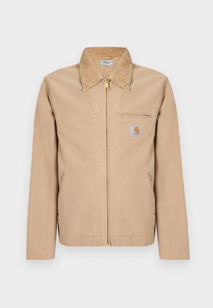 Beige jas met ritssluiting, spitse kraag, twee zijzakken, een borstzak met rits en een klein vierkant logo op de linkerborst.