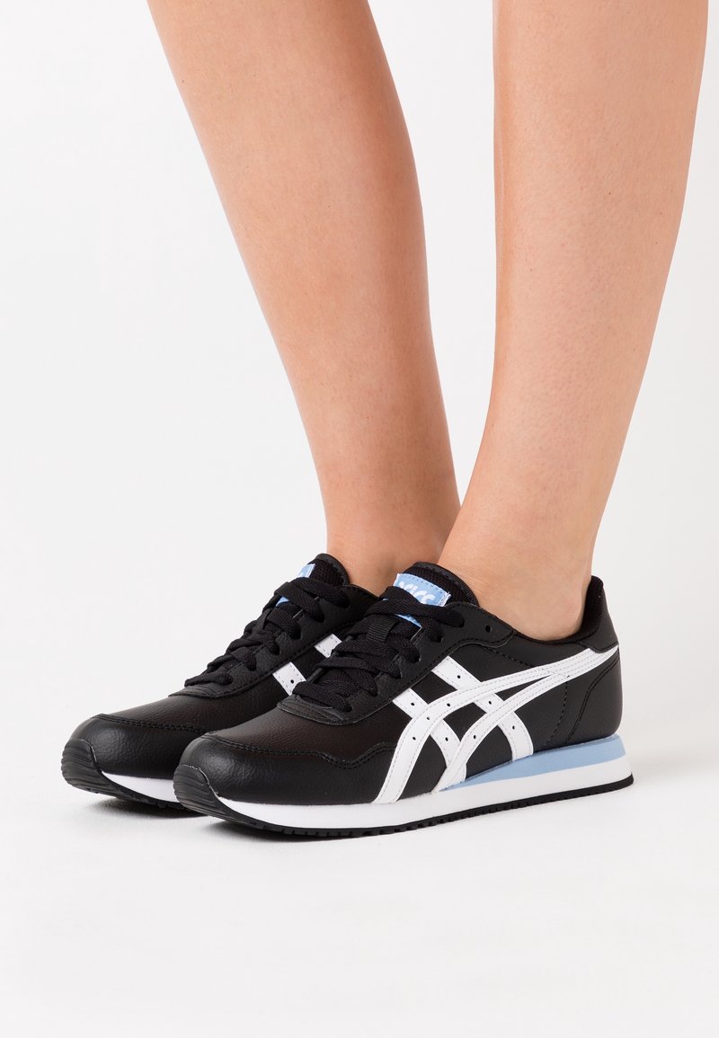 Chaussures de sport noires en cuir, dotées d'accents blancs et de trois bandes emblématiques. Semelle extérieure en caoutchouc avec un détail bleu au talon.