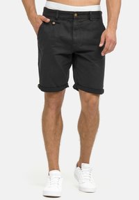Shorts en coton noirs à ourlets retroussés, avec une fermeture à bouton, deux poches avant et une ceinture blanche avec logo. Portés avec des baskets blanches.