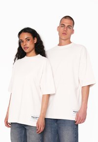 Hvide oversized t-shirts med korte ærmer, lavet af bomuld, med sidelabels og afslappet pasform, båret af to modeller mod en ensartet baggrund.