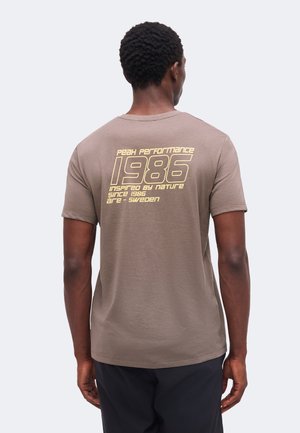 Uomo con capelli corti e ricci visto da dietro che indossa una maglietta marrone con testo giallo che recita "Peak Performance 1986 inspired by nature since 1986 Åre Sweden."