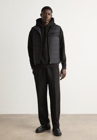 Black puffer vest over a black hoodie, paired with loose-fitting black trousers and chunky black shoes. Smooth texture, minimal design.
