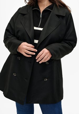Personne portant un manteau noir croisé sur un pull zippé rayé noir et blanc et un jean bleu, mains ajustant le devant du manteau.