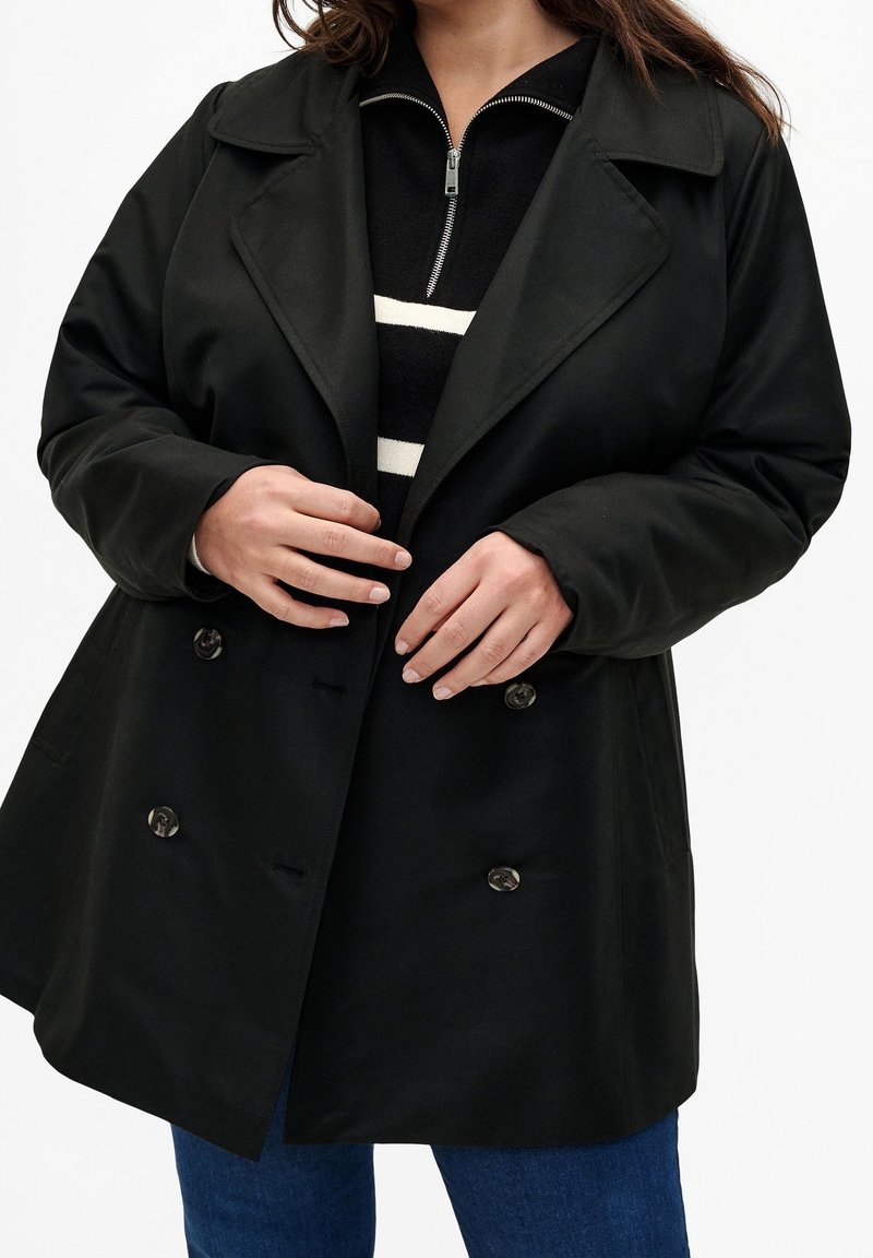 Personne portant un manteau noir croisé sur un pull zippé rayé noir et blanc et un jean bleu, mains ajustant le devant du manteau.