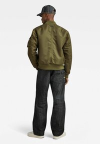 Veste bomber vert olive avec une finition satinée lisse, un ourlet côtelé et deux poches latérales, assortie à un pantalon en denim foncé et une casquette noire.