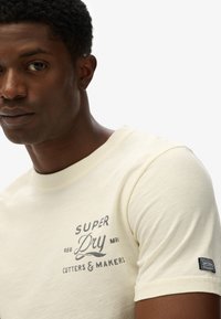 Camiseta de algodón color amarillo claro con una impresión en el pecho en gris oscuro que incluye el texto de la marca. Los detalles incluyen una etiqueta con el logo en la manga.