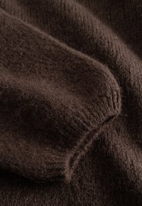 Rjavo pleteno oblačilo, ki prikazuje mehko, puhasto teksturo. Dizajn vključuje rebraste poudarke in gladko zaključne robove.