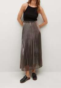 Zwarte mouwloze top gecombineerd met een geplooide, metallic midi-rok in grijstinten. Model draagt zwarte klompen met studs.
