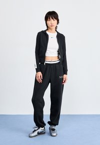 Svart zip-up hettegenser med hvite sidelister, hvit cropped topp, svarte joggebukser med logo, og sølv Nike-sneakers på en blå bakgrunn.