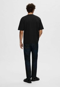 Tricou negru cu mânecă scurtă, cu o croială relaxată, purtat cu blugi negri și pantofi negri, fotografiat din spate pe un fundal simplu.
