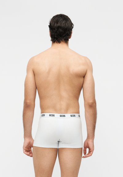 Homme aux cheveux foncés vu de dos portant un boxer blanc avec la ceinture logo "GCDS" sur un fond uni.