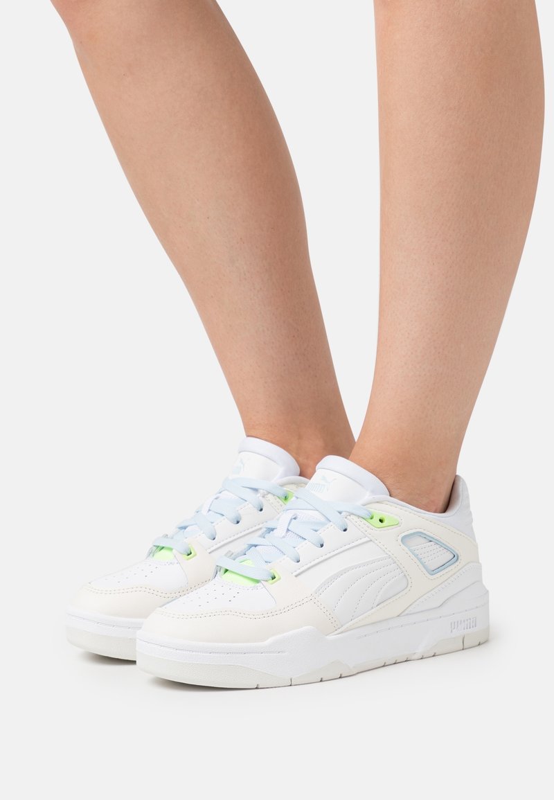 Baskets basses blanches Puma avec des touches vert clair portées sur des jambes nues sur un fond uni.