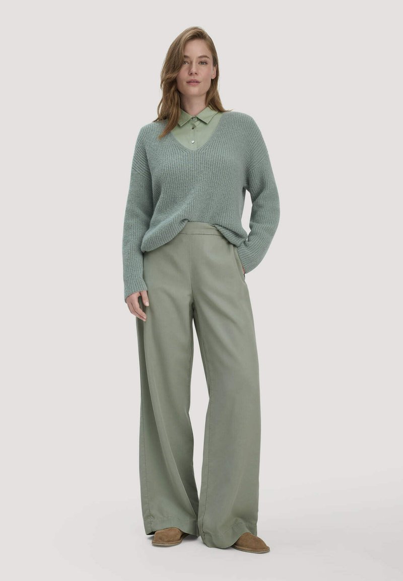 Femme portant un pull en tricot vert sauge par-dessus une chemise à col, un pantalon à jambes larges et des chaussures marron, debout devant un fond uni.