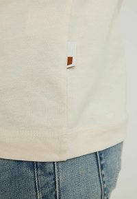 Hellbeige Baumwoll-T-Shirt mit einem seitlichen Etikett und braunem Akzent. Glatte Textur, kurze Ärmel und ein schlichtes Design.