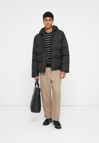 Giacca a vento nera con cappuccio, caratterizzata da un design trapuntato. Abbinata a una maglietta a righe e pantaloni beige. Il modello tiene una borsa nera.