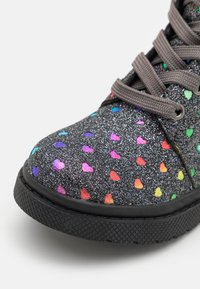 Zapatillas negras con purpurina que presentan patrones de corazones multicolores, superficie texturizada y cordones grises. Suela de goma negra con detalle de tracción.