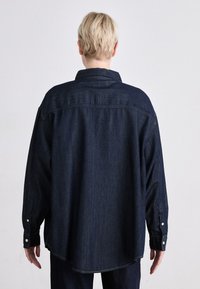 Mørkeblå denimskjorte med løs passform, knapp mansjetter og buet hem. Skjorten har klassisk krage og detalj med ryggstykke.