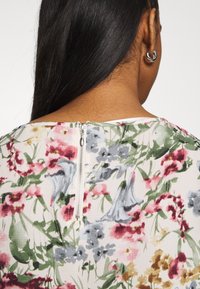 Vue de dos d'une personne aux longs cheveux foncés portant un haut à fleurs avec une fermeture éclair apparente et une boucle d'oreille en argent, sur un fond uni.