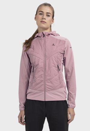 Frau trägt eine hellrosa Kapuzenjacke mit Reißverschluss und Reißverschlusstaschen, steht vor einem schlichten hellen Hintergrund.