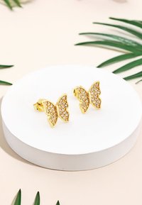 Guldörhängen i form av fjärilar med kristallaccenter, med intrikata detaljer och släta ytor, visade upp på en cirkulär vit pelare.