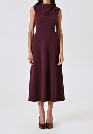 Robe midi sans manches de couleur bordeaux foncé avec un col drapé, portée avec des escarpins bordeaux assortis sur un fond blanc.