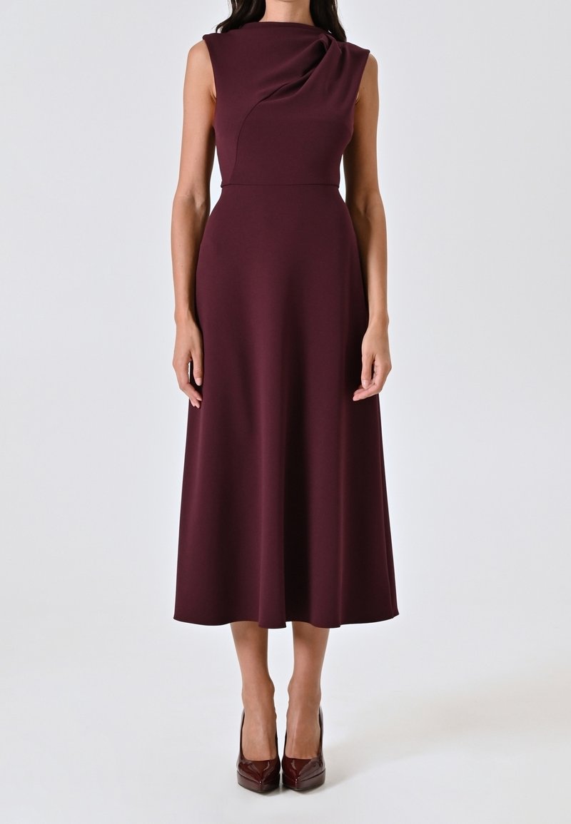 Robe midi sans manches de couleur bordeaux foncé avec un col drapé, portée avec des escarpins bordeaux assortis sur un fond blanc.