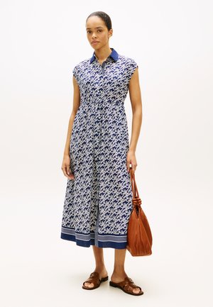Vrouw draagt een mouwloze midi-jurk met een blauw-wit patroon, houdt een bruine leren handtas vast en draagt bruine sandalen met open tenen.