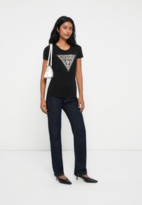 Donna che indossa una maglietta nera Guess, jeans blu scuro, tacchi neri e porta una borsa a tracolla bianca, in piedi contro uno sfondo bianco semplice.