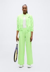 Γυναίκα που στέκεται όρθια κρατώντας ρακέτα τένις, φορώντας αθλητικό μπουφάν σε νέον πράσινο χρώμα και παντελόνι φαρδύ με λευκά αθλητικά παπούτσια.