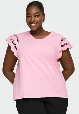 Usmievavá žena s tmavou pokožkou a vlasmi stáhnutými dozadu, oblečená v svetloružovom topu s čipkovanými volánmi na rukávoch a čiernych nohaviciach.