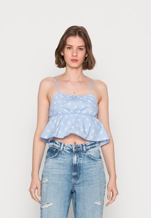 Top crop azul, sem mangas, com decote franzido e bainha ampla, apresentando estampa floral branca. Combinado com jeans de ganga rasgados de cintura alta.