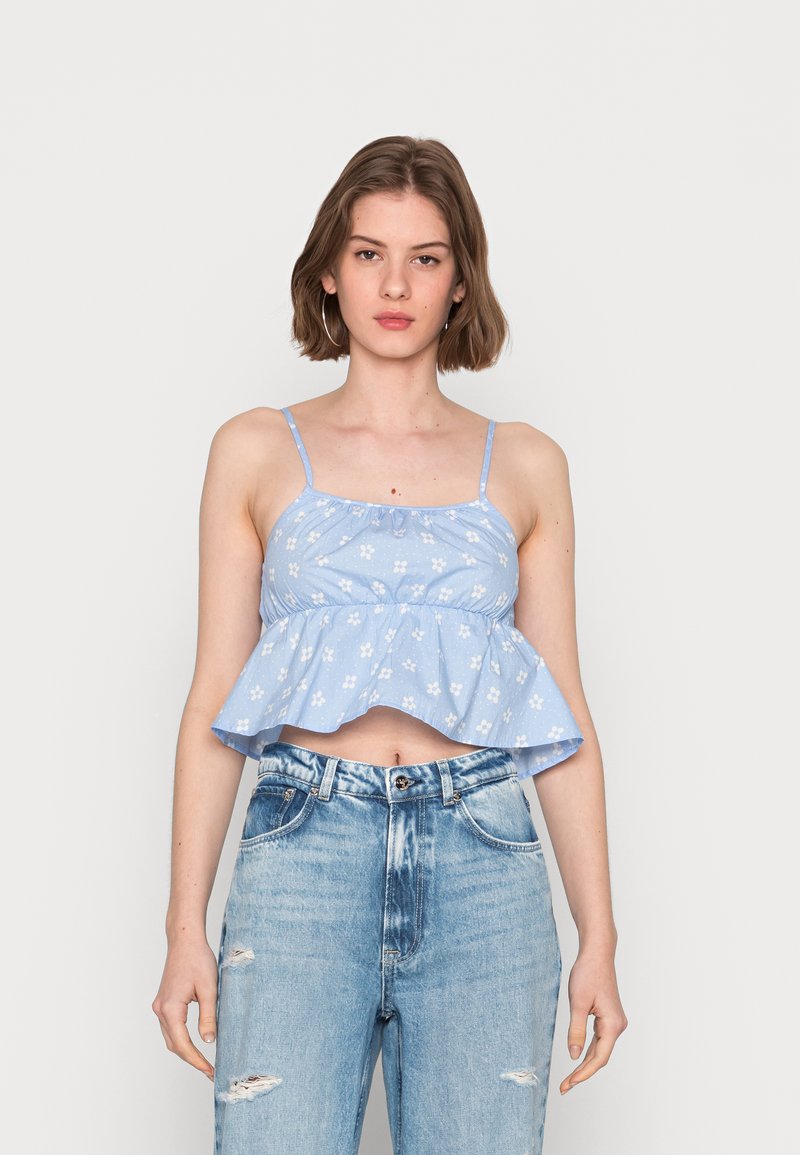 Top crop azul, sem mangas, com decote franzido e bainha ampla, apresentando estampa floral branca. Combinado com jeans de ganga rasgados de cintura alta.
