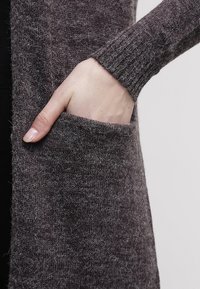 Main avec des ongles naturels placée à l'intérieur de la poche d'un vêtement en laine tricotée gris avec une manchette côtelée.