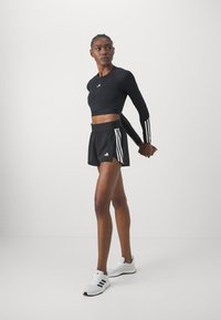 Crna dugorukava crop majica s bijelim Adidas logom, u kombinaciji s crnim šortsevima s bijelim bočnim trakama i bijelim Adidas tenisicama.