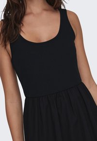 Musta ribattu hihaton paita, jossa on kaareva pääntie, ja se on varustettu valuvalle, kerääntyvälle hameelle. Materiaali näyttää olevan kevyttä ja pehmeää.