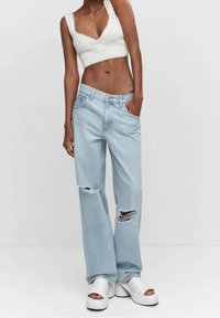 Jeans de tiro alto de color azul claro con rodilleras desgastadas, combinados con un top corto de punto blanco. La modelo está de pie con una mano en el bolsillo, llevándolo con sandalias de plataforma blancas.