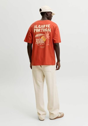 Homme portant un T-shirt rouge avec l'inscription « Algarve Portugal » et des motifs de plage dans le dos, pantalon crème, casquette beige et baskets, debout de dos.