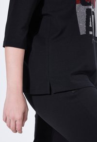 Černý top s krátkým rukávem a kulatým výstřihem, s bočními rozparky a mnohobarevným grafickým designem z kamínků na přední straně.