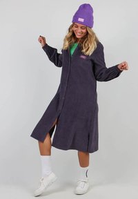 Robe chemise en velours côtelé violet avec un devant boutonné, des fentes sur les côtés et un patch logo. Associée à un bonnet en tricot violet et des baskets blanches.