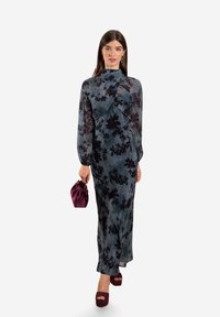 Langes, figurbetontes Kleid in dunkeltuerkis mit schwarzem Blumenmuster, seidige Textur, Hoher Kragen, lange Ärmel, kombiniert mit einer burgunderfarbenen Clutch und Plateau-Schuhen.