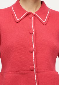 Κόκκινο πλεκτό ζακέτα με λευκή ραμμένη διακόσμηση κατά μήκος του γιακά και της γραμμής κουμπιών, με στρογγυλά κουμπιά καλυμμένα με ύφασμα.