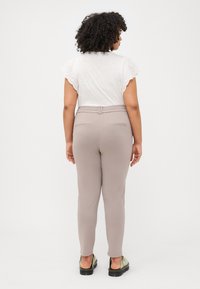 Eine Person mit lockigem Haar trägt ein weißes Oberteil und beigen Hosen und steht vor einem neutralen Hintergrund. Der Fokus liegt auf der Passform und dem Stil des Outfits.