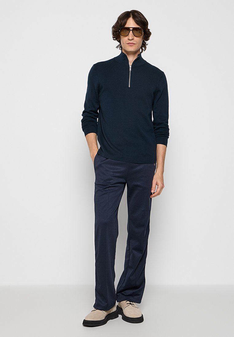 Marineblauwe pullover met een kwart-rits kraag, gecombineerd met donkere broek en lichtgekleurde schoenen. Model draagt een zonnebril en presenteert een casual outfit.