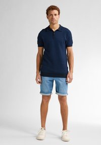 Námořnická modrá polo tričko s texturovaným vzorem, krátkými rukávy a límečkem, spárované s vyhrnutými světlými džíny a bílými teniskami.