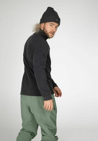 Maglione in pile nero con colletto alto, abbinato a pantaloni morbidi e larghi di colore verde. Il soggetto indossa un berretto scuro e strutturato e si trova in una posizione neutra.
